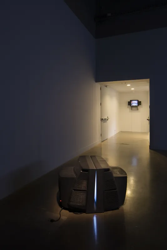 Two face-to-face monitors sit on the floor in a dark room, and monitor plays a video in a lighted area beyond.