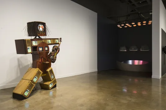 A figure made of cellos, color TVs, and antique TV cabinets, leans near a sculpture reflecting colored lights from above.