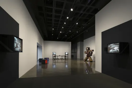 In a long view of a black and white gallery space, several video sculptures are displayed.