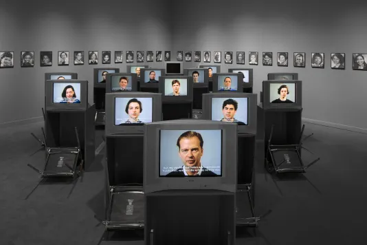 An array of video monitors on desks displaying faces, upside down chairs placed before each, portraits line the walls in back.  
