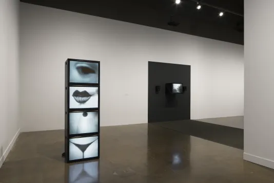 Totem of 4 monitors display videos of body parts, beyond which a monitor is mounted on a black section of a white wall.