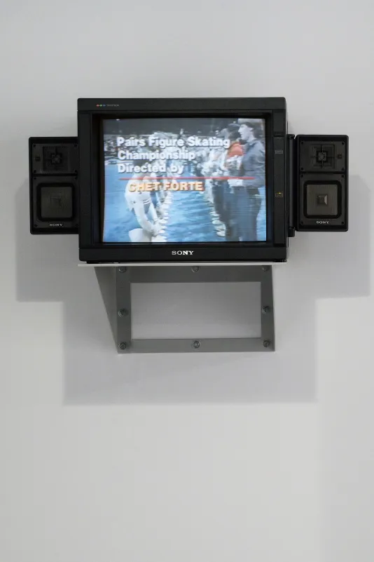 A mounted video monitor with attached speakers displays 2 rows of competition figure skaters, words superimposed over them.