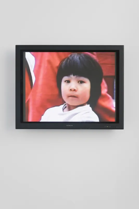 Video monitor shows a closeup of a young girl with short black hair staring at the viewer, background saturated in red.