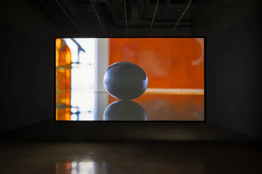 Video of black oval object with reflection, in a diffusely lit orange space, is projected on a hanging screen in a dark room.