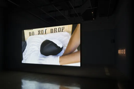 Video projection of a black object on a white pillow & 2 arms with gloved hands. Do Not Drop is stenciled on crate in back.