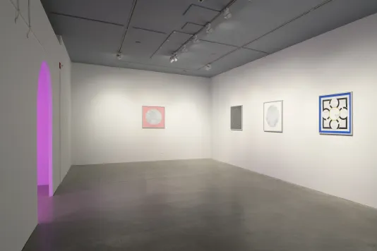 A pink light is seen through an arched doorway. Three artworks hang on the right wall, with a fourth hanging in the middle.