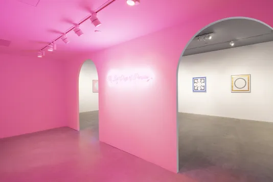 Pink light emanates from a neon sculpture and suffuses the space. Framed images are visible from two arched doorways.