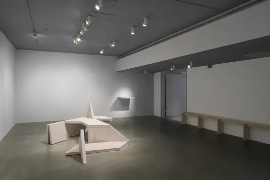 A concrete geometric bench sculpture sits on the floor, an object juts from a wall at back, a long bench lines the right wall.