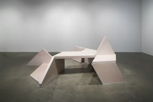 A concrete bench sculpture, made of geometric shapes with a triangle sticking up from the seat area, sits on a gray floor.