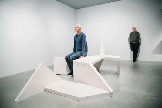 A white haired man sits up straight on a concrete bench sculpture, behind him a man with a white beard leans against a wall.