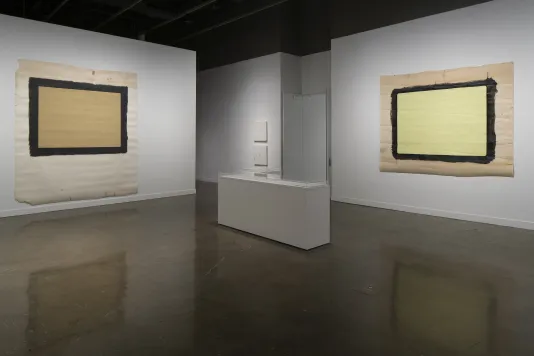 Two large pieces of paper with painted rectangles are mounted on perpendicular walls, with a glass-topped vitrine on the floor.
