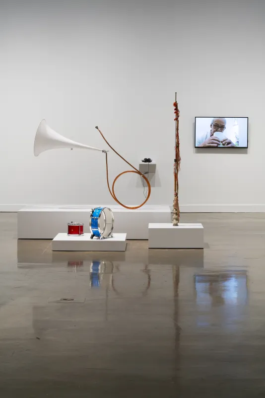 A collection of whimsical musical instruments are displayed on three white platforms. A video monitor displays a white male.