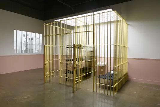 A yellow jail cell, with beds and a sink, stands in the middle of the space, while an image is projected on the wall to the left.