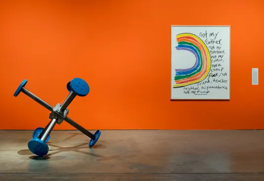 An orange gallery wall features artwork by Sable Elyse Smith, a crayon drawing of a rainbow, and an installation featuring blue buttons in the foreground.