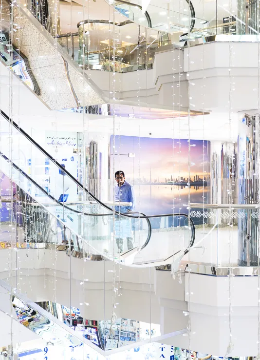 Next to a glass and mirrored escalator, a man stands before a picture of a pink gold sunset in a glossy 3-level mall atrium