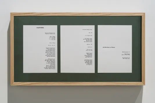 Hanging from a white wall, a wooden rectangle frame surrounds green matting and 3 pieces of paper on which addresses are shown.