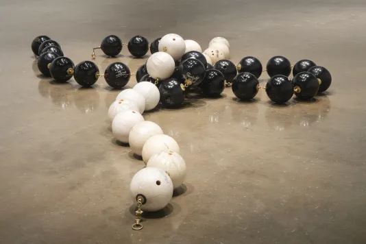 Black and white bowling balls are linked with brass hardware to create strands of monochromatic beads, white on top of black.