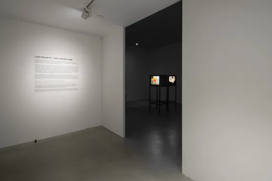 Two rooms, one illuminated and one in darkness, with text on the wall of the first room and videos on monitors in the other.