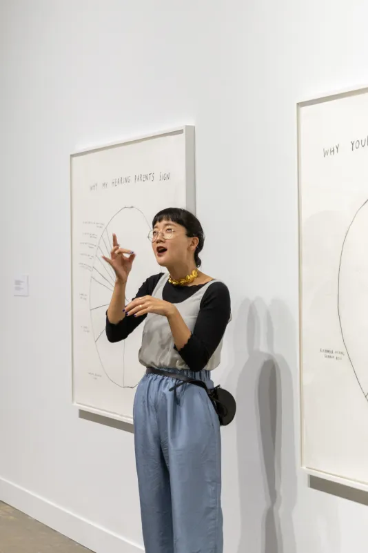 Artist, Christine Sun Kim signs at the exhibition opening in front of two framed pie-chart drawings on a gallery wall.