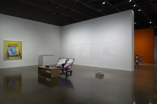 View of the List Center galleries with the "Mundane Futures" section of the exhibition. In the foreground is Aria Dean's video, in the background is a photo of hands holding a bedpan box with text "Futuro"