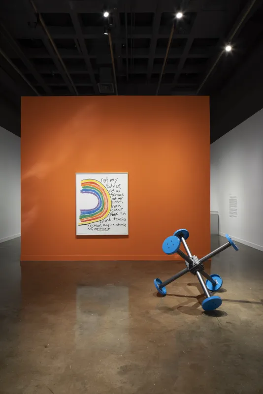 An orange wall with a large framed coloring book drawing of a rainbow on its side. In front are blue stools taken from a jail visiting center morphed into a structure that resembles a "jack" from the game "jacks."