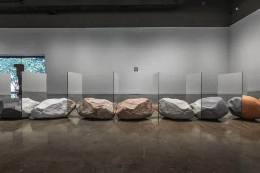 Installation view of objects resembling precious rocks are arraged in a straight row. Between each object is a mirror, creating the illusion that each rock is morphing into one another. They sit in front of a large glass window.