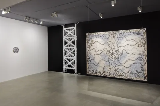 Painting with grayscale vines and the faint shadow of a pelvic bone hangs in front of a black wall from chainlink. To the left is a a small circular black and white painting with small illustrated figures. 