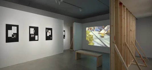 Projected color still of a playground and 4 paper works on black are shown on white walls with exposed wood framing.