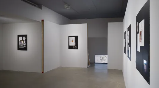 5 paper works on black displayed on white walls, a monitor on the floor sits between 2 constructed wall sections.