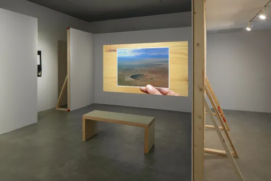 In a constructed alcove, projected still of hand holding a photo of a crater with distant blue mountains above a wood surface