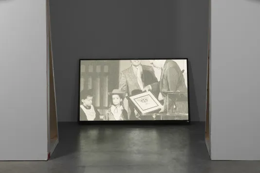A floor monitor shows a black-and-white still of 2 boys in cowboy costumes and 2 men in suits, 1 holds a framed certificate.