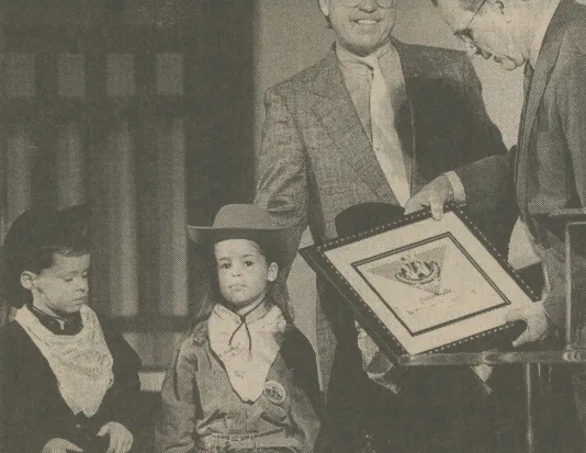 Black-and-white still of 2 men in suits, 1 about to present a framed certificate to 2 boys dressed as cowboys