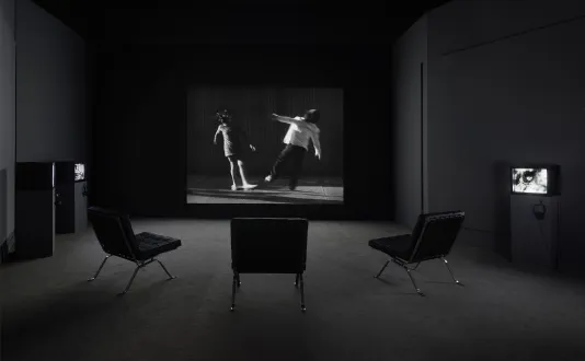 Photo of a black and white video projection and three chairs. Projection features a still of two children that appear to fall away from one another.