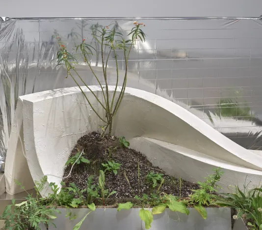 In a gallery at the List Center, an installation features a white plaster slumped figure resting between aluminum sheets, a mirror, and weeds.