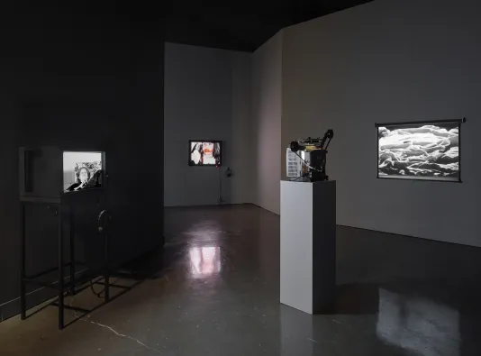 From left to right, a television set featuring a woman in black and white, a monitor featuring a young girl with lipstick smeared over her lips, and a projector and projection screen with black and white bedsheets.