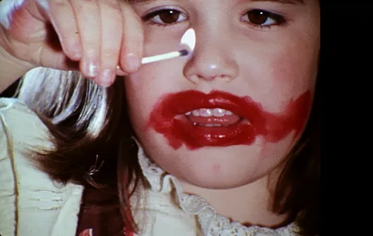 A child with light skin tone and brown eyes holds a lit match; her mouth, teeth, and face are smeared with red lipstick.