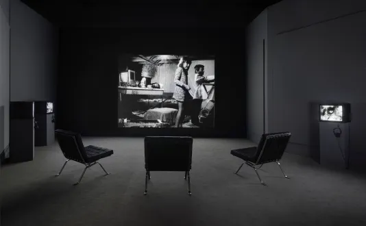 View from the back of the gallery with three large leather seats facing a black and white projection of two children playing in a messy room.