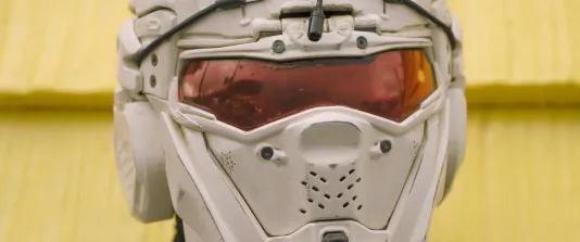 extreme close up of a person wearing a futuristic storm-trooper-like helmet posing  in front of  bright yellow vinyl siding.