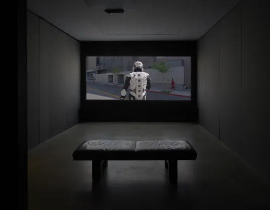 A dimly lit gallery space includes two video screens, one closer to the forefront on the left and one in the back right wall. A projector and a bench are also shown.