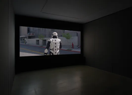 A dimly lit gallery space includes a screening of a storm tropper with its back turned to the screen and a soft bench. 