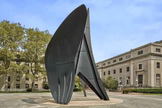 Large steel sculpture in a courtyard with trees and buildings in the background.