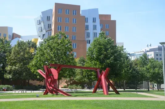 Red steel sculpture on a field of grass with trees and buildings in the background.