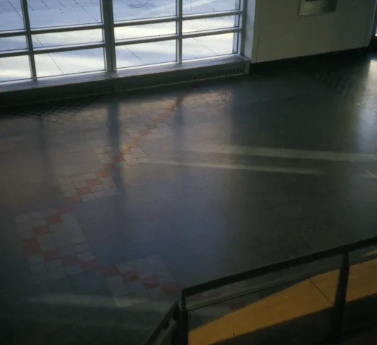 Grey tiled floor with red tiles in 2 diagonal lines lit by natural light from the windows at the top of the frame.