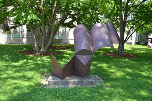 Steel sculpture on a lawn featuring several hollow segments and two adjacent triangular prisms that stand on end in contact with the ground.