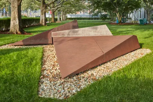 Corten steel sculpture surrounded by pebbles on a lush green grass.