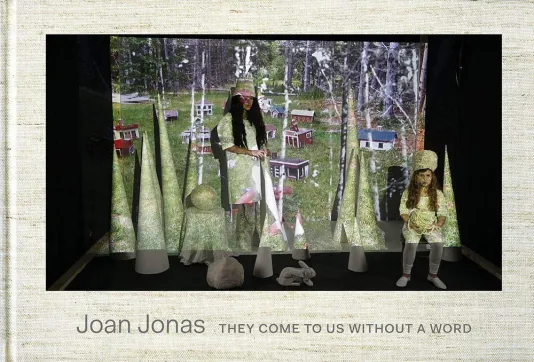 Book cover for Joan Jonas: They Come to Us without a Word featuring a large projector with an image of small houses and two females standing in front of the screen dressed in all white. There are tall cone sculptures around the two females and a sculpture of a rabbit.