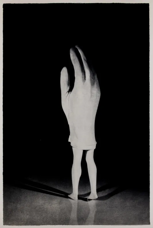 Black and white photograph of a person standing wearing a giant glove that covers their head to their lower thighs.