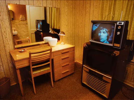 Corner of a motel room with a desk on the left and a large mirror above and a tv on a tall stand to the right