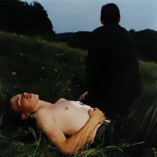 A man lies in a field of grass shirtless while a dark figure walks away to his left.