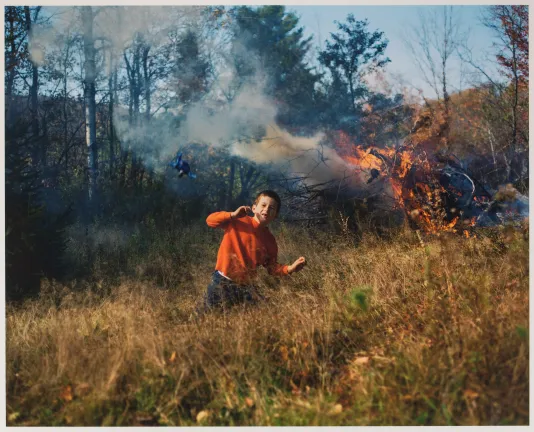 Photograph of a young boy in red sweater standing in a field white the woods behind him is on fire.
